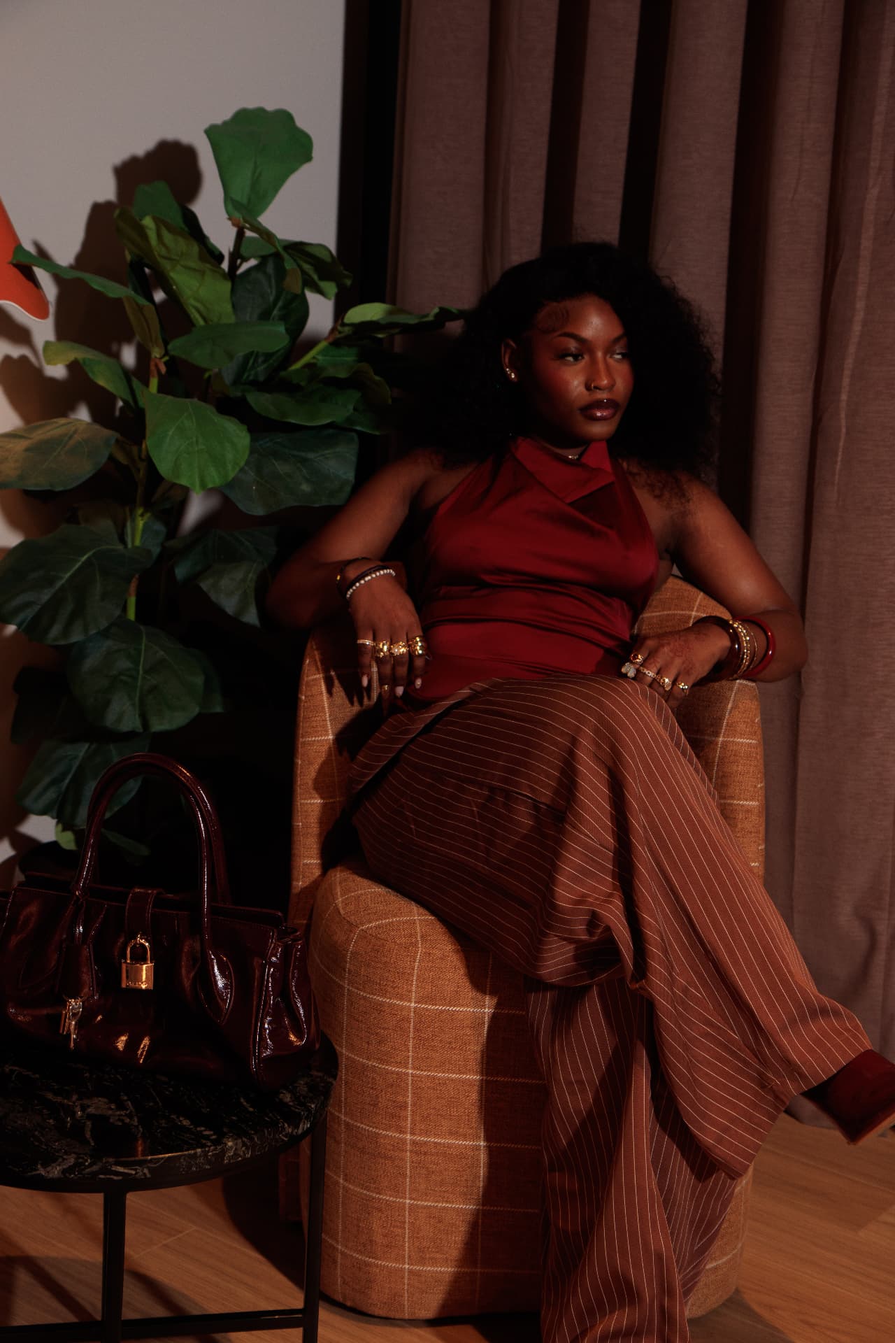 Editorial portrait — woman in a rust-red top and pinstripe trousers seated in an armchair
