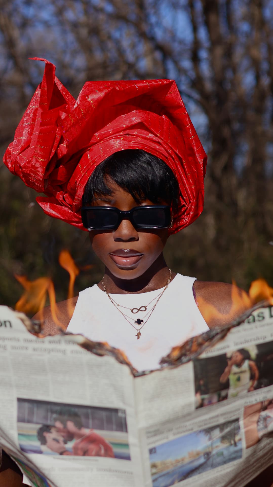 Portrait of a woman in a tall red headwrap and sunglasses holding a burning newspaper across her chest