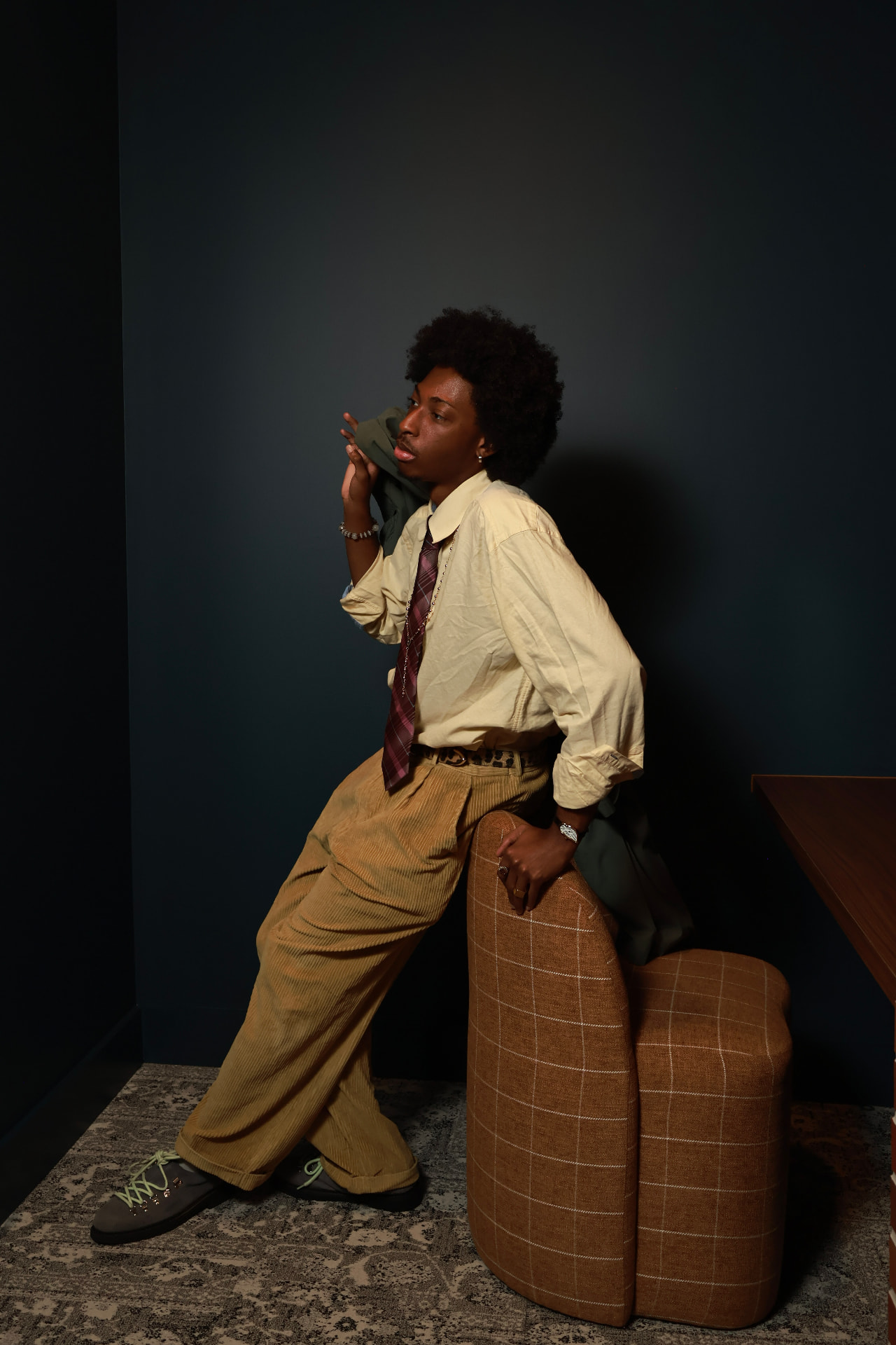 Editorial portrait — man in a cream oversized shirt, plaid tie and tan corduroy trousers leaning against a windowpane-plaid chair